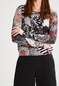 Person med långt brunt hår bär en långärmad topp med svarta, vita och röda blommönster och svarta byxor, med armarna korsade vid midjan.