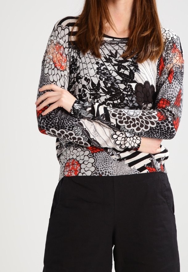 Person med långt brunt hår bär en långärmad topp med svarta, vita och röda blommönster och svarta byxor, med armarna korsade vid midjan.