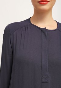 Blouse bleu marine en tissu lisse avec un col rond, des détails froncés aux épaules et une fermeture carrée à boutons à l'avant.