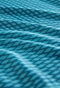 Textilväv i teal med ett diamantmönster. Materialet är slätt och visar mjuka veck och en lätt lyster över ytan.