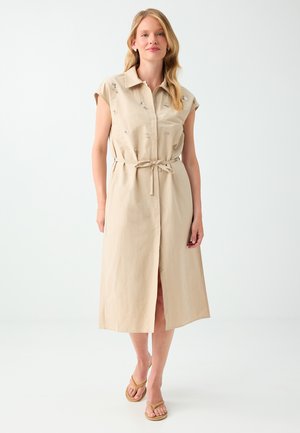 Vrouw die een beige mouwloze midijurk draagt met een kraag, bloemr embroidery en een gestrikte tailleband, gecombineerd met beige sandalen.