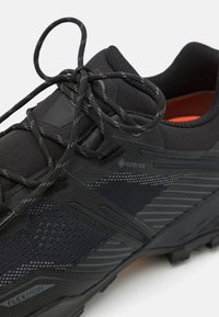 Musta urheilukenkä, jossa on teksturoitu yläosa, Gore-Tex -brändäys ja kestävä kumipohja. Varustettu turvallisella nauhoitusjärjestelmällä ja vahvistetuilla yksityiskohdilla.