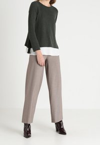 Haut à manches longues vert foncé avec des fentes latérales sur une couche blanche, associé à un pantalon large à motif pied-de-poule beige et des bottes brunes brillantes.