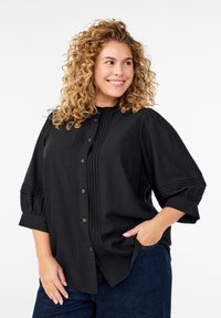 Svart blus med veckad framstycke, rund halsringning och knappstängning. Ärmarna är samlade och har manschettdetaljer.