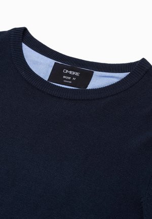 Pull-over tricoté bleu marine avec un col rond, présentant un accent bleu à l'intérieur du col. L'étiquette indique "OMBRAE" et la taille "M".