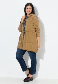 Giacca con cappuccio marrone realizzata in tessuto morbido, caratterizzata da cuciture arricciate. Abbinata a jeans blu e mocassini scuri, creando un look casual.