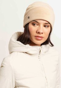 Beanie in pile beige con una texture liscia, caratterizzato da un piccolo logo in evidenza. Abbinato a una giacca puffer bianca con cappuccio.