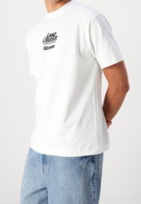 Biała bawełniana koszulka z krótkim rękawem, z czarnym nadrukiem graficznym na piersi. Krój luźny i lekko za duży.