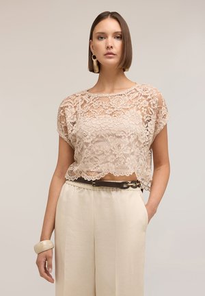 Frau mit kurzen braunen Haaren, die ein beiges Spitzentop, cremefarbene weit geschnittene Hosen, einen dunklen Gürtel, große Ohrringe und ein massives Armband trägt.