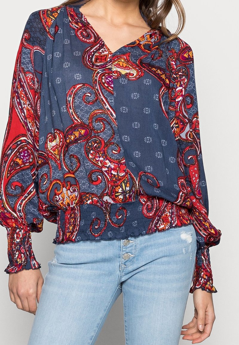 Blusa vibrante de color azul y rojo con un escote en V, mangas largas y dobladillo fruncido. Presenta intrincados giros y diseños florales.