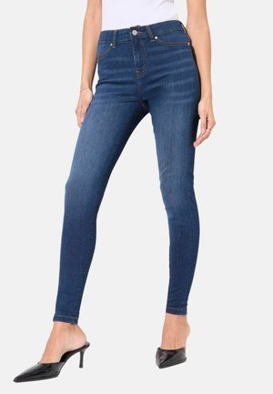 Jeans en denim bleu taille haute avec une coupe slim, présentant une fermeture à boutons, des poches avant et des coutures contrastées. Assortis avec des mules noires.