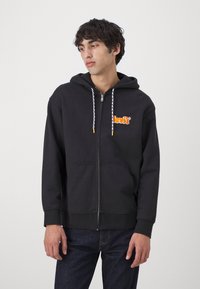Sudadera con cremallera negra de algodón con una textura suave, que cuenta con bolsillos frontales, capucha ajustable con cordón y un logo naranja en el pecho.