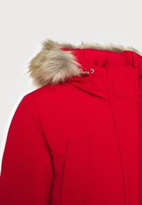 Chaqueta parka roja con capucha forrada de piel, que cuenta con cierre de cremallera, bolsillo frontal y tejido texturizado para aislamiento.