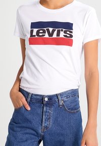 Bílý tričko s krátkým rukávem s centrálním červeným a modrým logem Levi's. Spárované s džínami s vysokým pasem modré barvy, které mají opotřebovaný vzhled.