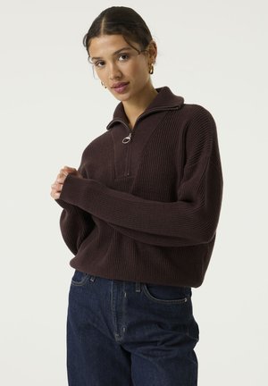 Woman wearing a dark brown ribbed zip-neck sweater and dark blue jeans, standing against a plain light background.