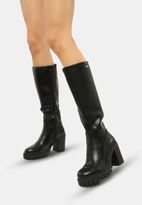 Botas negras de cuero liso hasta la rodilla, con un tacón ancho, suela de goma texturizada y una cremallera lateral para facilitar su uso.