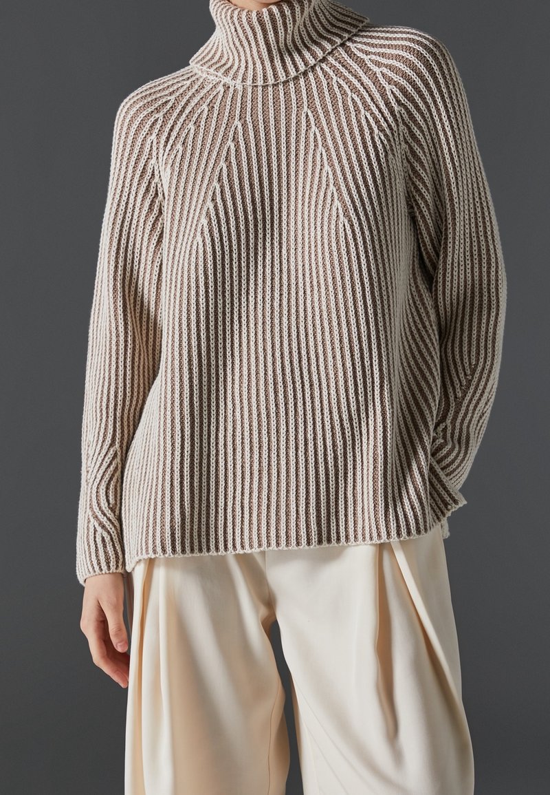 Personne portant un pull à col roulé côtelé rayé beige et blanc et un pantalon ample de couleur crème sur un fond gris foncé.