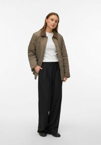 Veste extérieure matelassée marron avec un col marron, portée sur un pull blanc et un pantalon noir à jambes larges. Fermetures à pression.