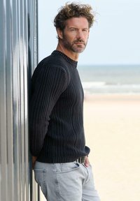 Schwarzer, gerippter Pullover mit rundem Ausschnitt, getragen zu hellgrauen Jeans. Im Hintergrund sind ein Strand und der Ozean zu sehen, daneben eine metallische Wand.