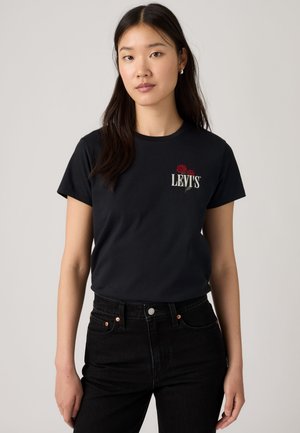 Femme aux longs cheveux foncés portant un t-shirt noir Levi's avec un logo de rose rouge et un jean noir taille haute, debout devant un fond uni.