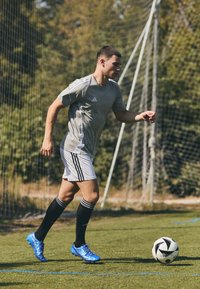 Athlet in grauem kurzärmeligen Trikot und weißen Shorts mit schwarzen Streifen, trägt schwarze Socken und blaue Fußballschuhe, dribbelt einen Fußball auf Gras.