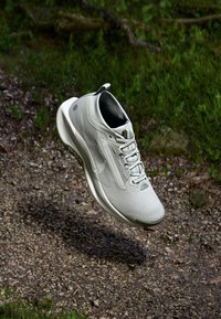 Light gray athletic shoe with a breathable mesh upper, textured accents, and a cushioned sole, displayed mid-air above a gravel surface.