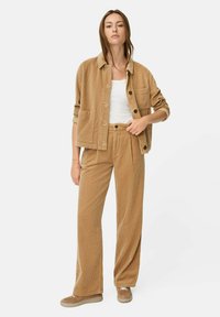 Beige corduroy jasje met bijpassende broek; knoopsluiting, textuurstof, zakken op het jasje, gecombineerd met een wit topje en beige schoenen.