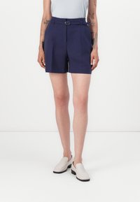 Marinblå shorts har en bältesmidja, skräddarsydd design med veck och är i medellång längd till mitten av låret, i kombination med ljusa slip-on skor.