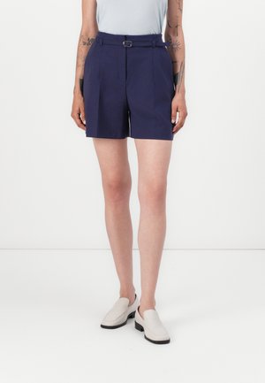 Marineblå shorts har en belted waist, skreddersydd design med folder, og en mid-thigh lengde, kombinert med lyse slip-on sko.