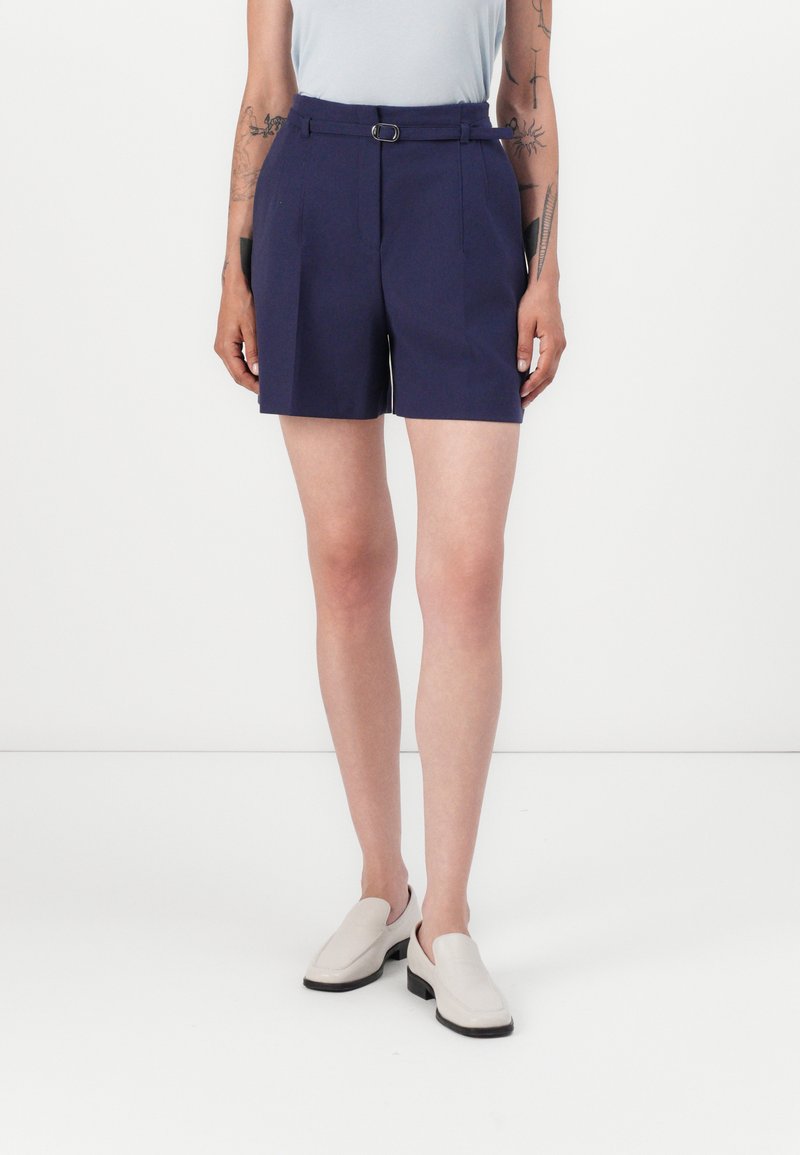 Marinblå shorts har en bältesmidja, skräddarsydd design med veck och är i medellång längd till mitten av låret, i kombination med ljusa slip-on skor.