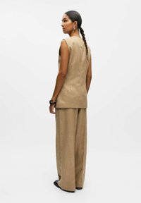Gilet beige ajusté et pantalon large en tissu texturé, avec des lignes épurées et un design minimaliste. Accentué par des chaussures noires élégantes.