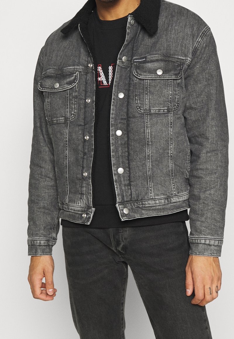 Chaqueta de mezclilla en negro desgastado con un acabado texturizado, cierre de botones, bolsillos en el pecho y un suave cuello negro. Lleva puesta sobre una camisa negra.