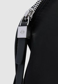 Bolso negro de nailon con cremallera plateada, con una correa de cuero suave y un adorno metálico con un detalle de logotipo cuadrado.