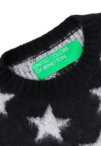 Svart stickad tröja med vita stjärnmönster, ribbad krage och en grön etikett som står "UNITED COLORS OF BENETTON." Mjuk textur.