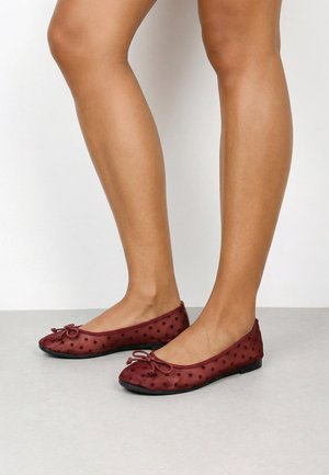 Piernas con bailarinas burdeos transparentes con lunares, pequeño detalle de lazo y suelas negras sobre un suelo gris claro frente a una pared blanca.