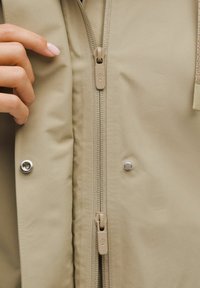 Béžová bunda s dvojitým zipem, patentními knoflíky a hladkou texturou. Na táhle zipu je detail s logem, což zvýrazňuje design.