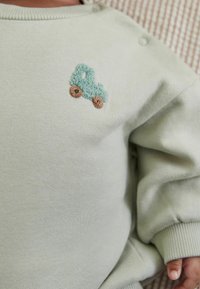 Jasnozielona bluza z fakturowanym, haftowanym wzorem niebieskiego samochodu ciężarowego oraz brązowymi kołami, z prążkowanymi mankietami i zatrzaskami na ramionach.
