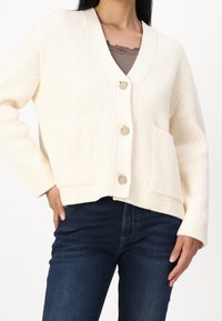 Cremfarbener gerippter Cardigan mit V-Ausschnitt, drei Knöpfen und Taschen auf der Vorderseite, der ein strukturiertes Strickmuster und einen lockeren Schnitt aufweist.