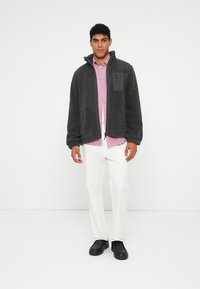 Pelēka vilnas jaka ar augstu apkakli un rāvējslēdzēju priekšpusē, ar melnu krūšu kabatu. Valkāta virs gaiši rozā krekla un baltām biksēm.