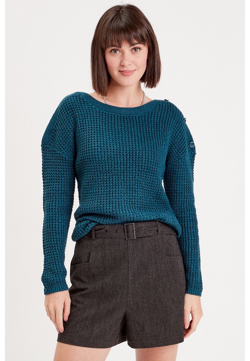 Pull en maille vert d'eau avec des manches longues, texture côtelée, encolure large et détail de boutons à l'épaule, associé à un short gris.