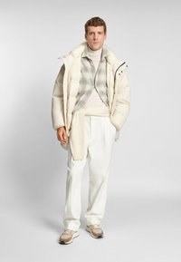 Giubbotto imbottito beige chiaro, indossato sopra una camicia a quadri e un dolcevita. Pantaloni bianchi a gamba larga e sneakers beige completano il look.