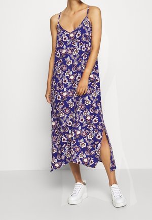 Femme portant une robe midi fleurie violette sans manches avec une fente latérale et des baskets compensées blanches, debout devant un fond gris clair.