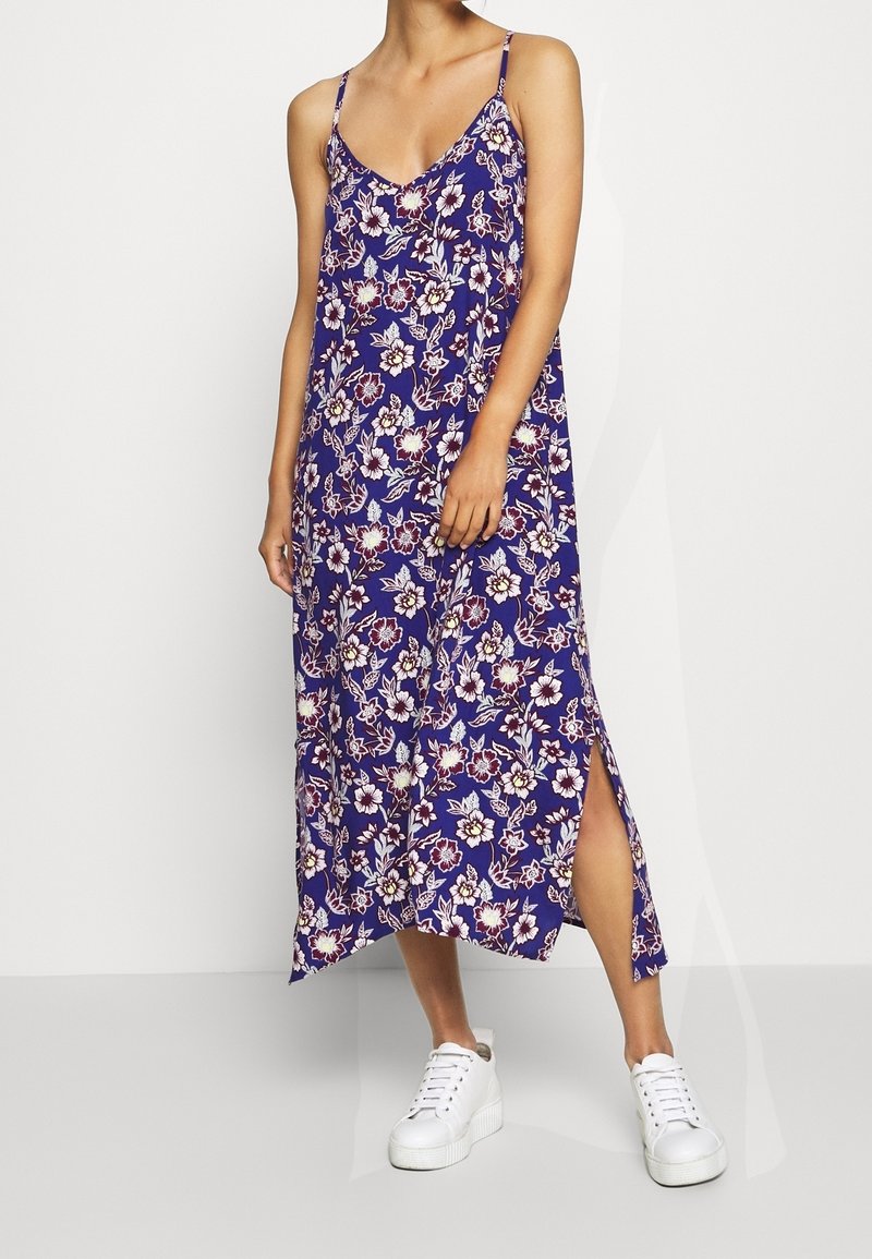 Femme portant une robe midi fleurie violette sans manches avec une fente latérale et des baskets compensées blanches, debout devant un fond gris clair.
