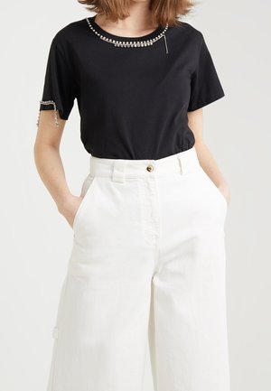 Kvinde iført en sort top med korte ærmer og rhinstenkant på hals og ærmer, kombineret med højtaljede, vidtbenede, hvide bukser, hænderne i lommerne.