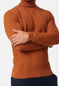 Homme portant un pull à col roulé orange rouille avec un motif en chevrons en tricot et un pantalon bleu marine, les bras croisés sur la poitrine.