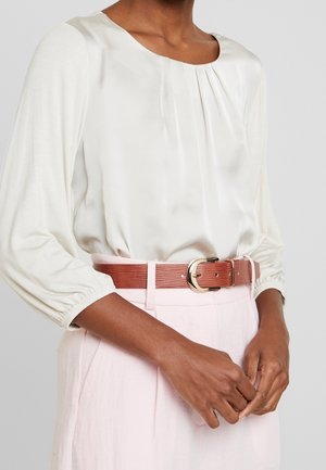 Vrouw die een witte blouse met lange mouwen en plooien draagt, ingestopt in lichtroze broek die wordt vastgehouden door een bruine riem met textuur en een gouden gesp.