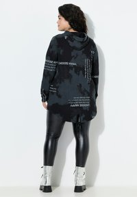Sudadera negra con capucha, decorada con patrones florales y texto blanco. Mangas largas, con la parte trasera alargada, combinada con legging negros brillantes y botas tobilleras plateadas.