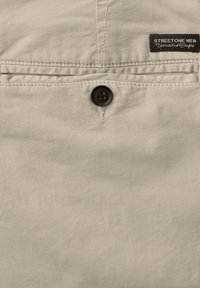 Tissu beige avec une poche arrière dotée d'une fermeture à bouton et d'une étiquette noire "STREET ONE MEN Unmatched Comfort" en haut à droite.