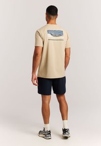 Beige T-shirt met korte mouwen met een afbeelding van een berg en zon op de achterkant, gecombineerd met marineblauwe korte broek en sportschoenen.