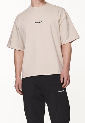 Beige T-Shirt mit kurzen Ärmeln und Rundhalsausschnitt, mit einem kleinen schwarzen "PEGADOR"-Logo auf der Brust. Hergestellt aus weichem Stoff, entspannter Schnitt.